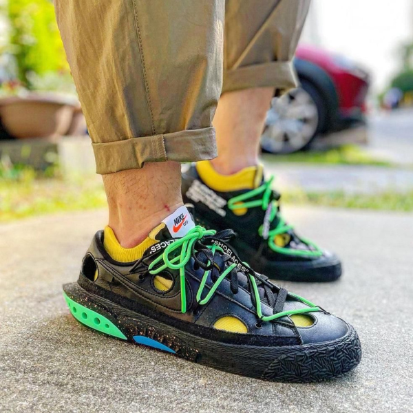 Close-up of black Nike sneakers with neon green laces and yellow socks, rolled khaki pants, standing on pavement.