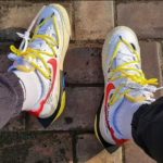 Top-down view of white sneakers with bright yellow laces on brick pavement.