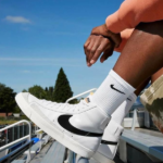 White Nike high-top sneakers with a black swoosh, worn with white socks, resting on bleachers outdoors on a sunny day.