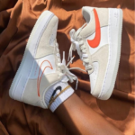 Pair of white Nike sneakers with orange swoosh and beige suede panels, laces tied, resting on a brown fabric surface; person wearing white socks with black and gold stripes.