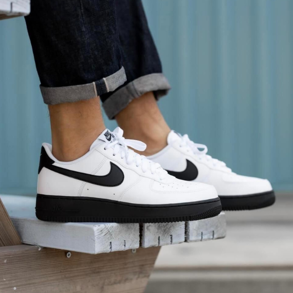 Pair of white Nike sneakers with black swoosh on a person standing on a weathered wooden bench, jeans rolled at the cuffs.