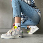 Person crouching and tying light gray Nike sneakers with pink laces, white socks, and light-wash jeans in a casual indoor setting
