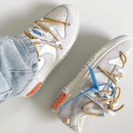 Pair of white and gray sneakers with blue and tan laces on a light floor, worn with jeans.