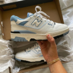 White and light-blue New Balance 550 sneakers in a cardboard box, with one shoe held by a hand showing the side detail and branding.