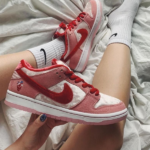 Hand holding a pink and red Nike sneaker while two similar sneakers rest on a bed with white sheets and socks visible in the background.