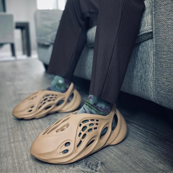 Person wearing tan, ventilated slip-on clogs with patterned socks indoors beside a sofa and hardwood floor.
