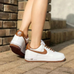 White Nike Air sneakers with brown accents and swoosh, worn by a person standing on a brick walkway near steps.