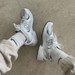 Untitleddesign-2022-09-29T142228.270_a6aea469-bf22-4c23-aa2e-9d79caa42407 | UAE SPORT STORE Two white New Balance sneakers worn with light socks and rolled-up pants, resting on a concrete floor.