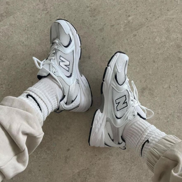 Two white New Balance sneakers worn with light socks and rolled-up pants, resting on a concrete floor.