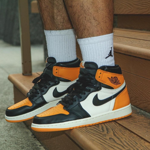 Person wearing orange, black, and white Air Jordan 1 sneakers standing on wooden steps with white crew socks visible