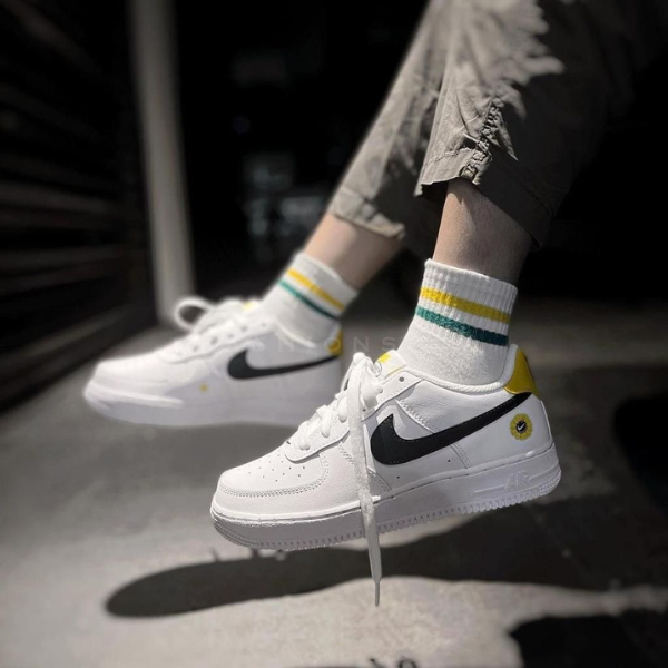 White Nike sneakers with black swoosh and yellow heel tab, worn with socks that have green and yellow stripes, shown mid-air as the person walks or jumps.