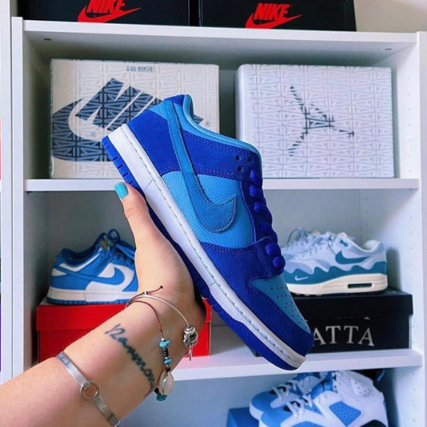 A hand holds a blue Nike sneaker in front of shelves with other sneakers and shoe boxes behind it.