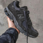 Hand holding a black suede New Balance running shoe with a thick dark sole against a gray concrete background.