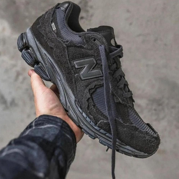 Hand holding a black suede New Balance running shoe with a thick dark sole against a gray concrete background.
