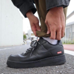Person tying the laces of a black Nike sneaker with a small Supreme logo on the side, on a city sidewalk.