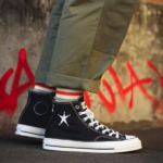 Close-up of black high-top sneakers with a white star, white laces, and white sole, worn with rolled olive pants.