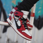 Red, white, and black Air Jordan 1 sneakers worn on a hanging leg, with a graffiti background.