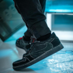 Close-up of a black suede sneaker with white stitching, worn by a person stepping forward on a blue-lit floor.