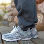 Untitleddesign-2023-01-05T122629.494_e05f980a-dad6-457d-9eb7-26026b92449a | UAE SPORT STORE Gray New Balance running shoes with white laces on a person standing on a wooden surface, with rocks in the background.
