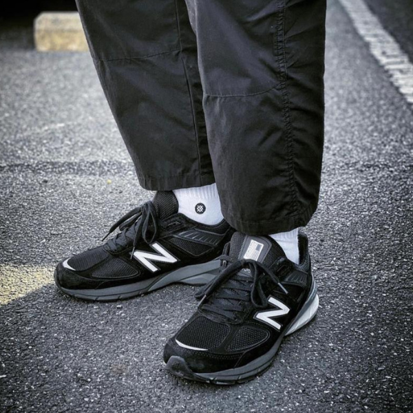 Pair of black New Balance sneakers with white socks standing on a street or parking lot surface.