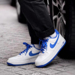 White Nike sneakers with blue swoosh and laces on a cobblestone sidewalk beside a car wheel