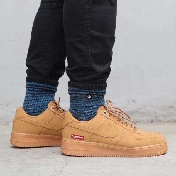 Person wearing tan Nike Air Force 1 sneakers with a red Supreme logo, paired with blue knit socks and black pants, standing on a concrete floor.