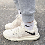 Beige mesh Nike running shoes with a black swoosh, worn with white socks and gray joggers on pavement.
