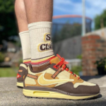 Close-up of tan and brown sneakers with orange laces worn with cream socks on a concrete surface.