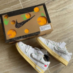 White sneakers with pale yellow soles and light green laces placed beside a brown shoebox with a black Nike swoosh and orange sticker circles on a tiled floor.