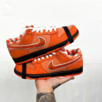 Pair of bright orange Nike sneakers with white laces and a black midsole stacked on a hand against a white background, showing the swoosh logo.