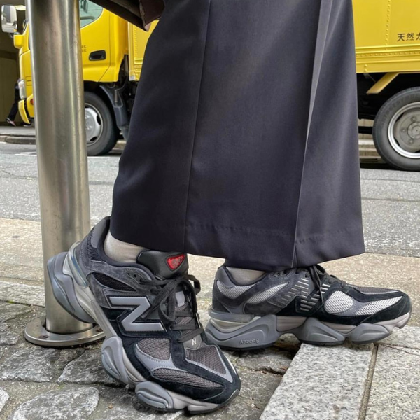Person wearing gray-and-black chunky sneakers and dark wide-leg pants, standing on a sidewalk beside a metal pole with yellow trucks in the background.
