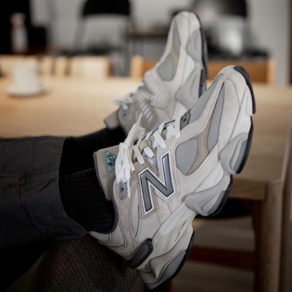 Untitleddesign-2023-02-09T122042.767_c8f1d260-6ac1-4ab6-a901-c09f6594a0a8 Pair of white and gray New Balance sneakers resting on someone’s feet in an indoor casual setting with a wooden table in the background.