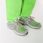 Person wearing gray-green New Balance sneakers with lime-green pants standing on a white surface.