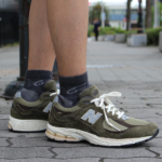 Person wearing olive-green New Balance sneakers with white laces standing on a paved sidewalk outdoors.