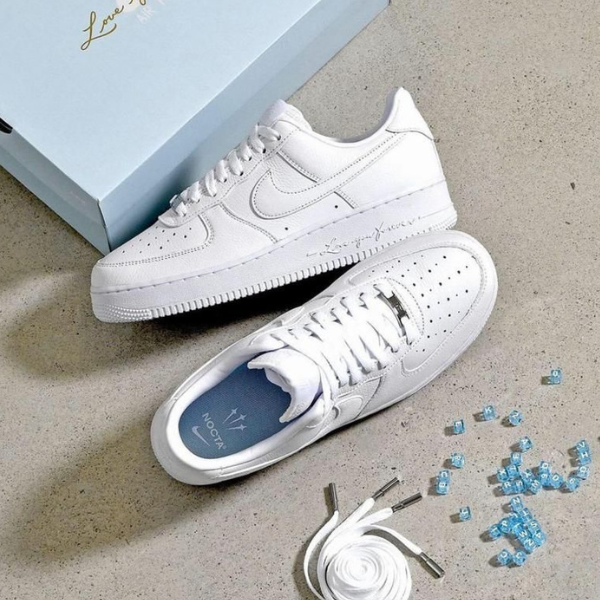 White sneakers with perforated toe cap placed on a light gray floor next to a pale blue shoebox; laces untied, blue insole visible, and small blue decorative beads with a coiled white shoelace nearby.