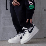 Close-up of white Nike Air Force 1 sneakers with black swooshes, worn with black jeans against a gray concrete wall.