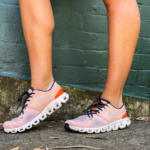 Pair of pink athletic sneakers with orange heel accent and black laces, standing on a sidewalk in front of a green brick wall.