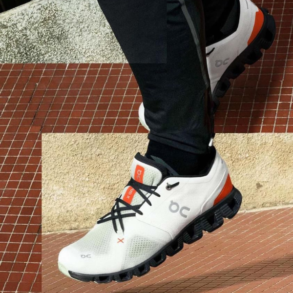 White athletic running shoe with black laces and orange details, stepping up a red-tiled staircase; visible OC logo on the side.