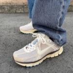 Pair of beige and gray running shoes with white laces on a person wearing blue jeans standing on a paved surface, outdoors on asphalt.