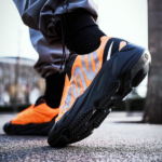 Close-up of an orange running shoe with a black sole on a wet pavement, a runner tying the laces shown from the side.