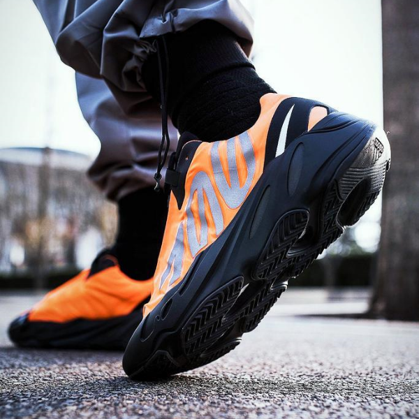 Close-up of an orange running shoe with a black sole on a wet pavement, a runner tying the laces shown from the side.