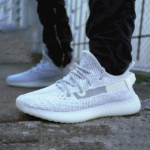 Glittery white knit sneakers on a person standing on a concrete surface, wearing black pants, side view showcasing the shoe's textured upper and thick sole.