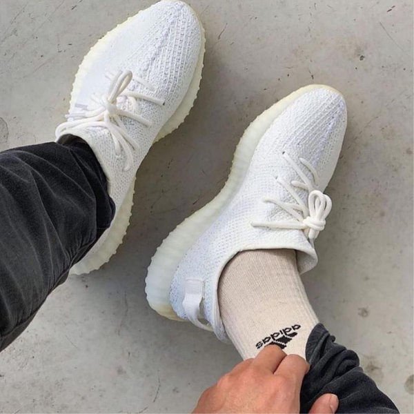 Top-down view of white knit sneakers with chunky off-white soles, paired with beige Adidas-branded socks on a gray floor.