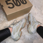 Pair of light gray Yeezy 500 sneakers with beige soles on a white shag rug next to a cardboard box labeled '500'.