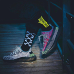 Pair of sporty sneakers on a wooden deck, one foot in a black sock with white arrows and a yellow taped label nearby.
