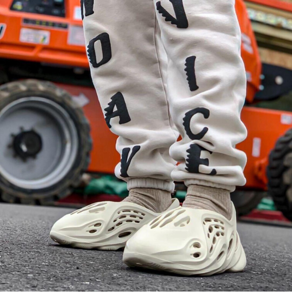 Person wearing light gray joggers with black scattered letters, standing in white sneakers on pavement, orange machinery in the background.