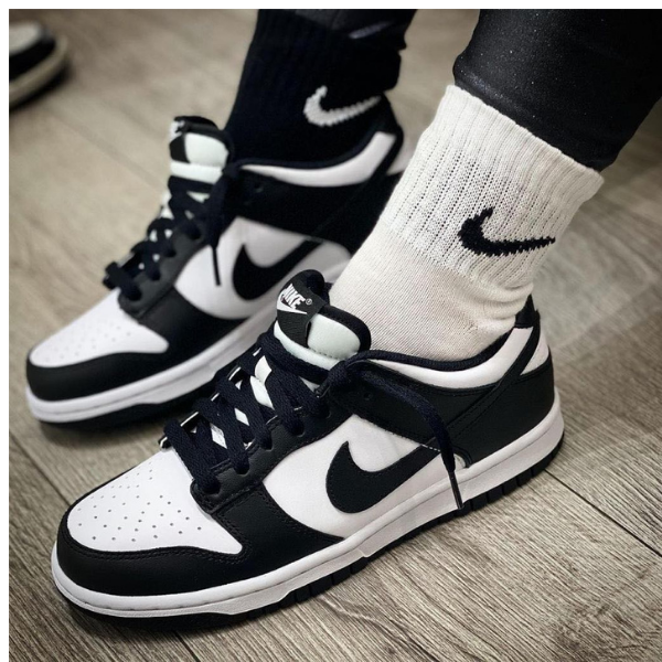 Close-up of black and white Nike sneakers on the feet, with white socks showing the swoosh, standing on a wooden floor.