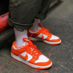 Close-up of a person wearing white crew socks and orange-and-white Nike sneakers standing on a dark pavement.