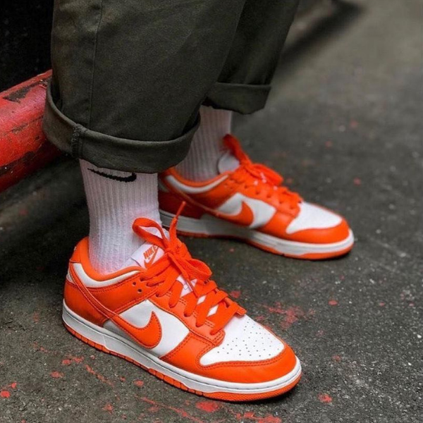 Untitleddesign_52_daf427c0-da5a-43fd-abe8-4485ed4e1c9b Close-up of a person wearing white crew socks and orange-and-white Nike sneakers standing on a dark pavement.