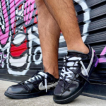 Pair of black sneakers with white swoosh worn by a person standing on concrete in front of a colorful graffiti mural on a metal shutter.